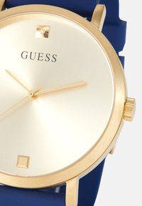 Montre Guess dorée avec cadran crème, index horaires carrés minimalistes et bracelet bleu, présentée en gros plan depuis le côté droit.