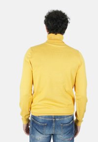 Maglione giallo a collo alto in tessuto morbido, con collo e polsini a coste, abbinato a jeans di denim blu, mostrato di spalle.