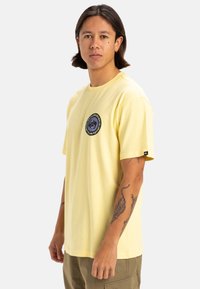 Camiseta de algodón amarilla con mangas cortas, escote redondo y un logotipo gráfico circular en el pecho. Presenta un corte relajado y está sin adornos.