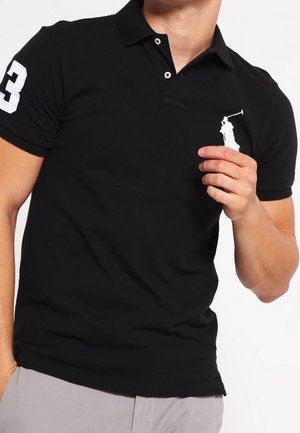 Mężczyzna noszący czarną koszulkę polo z białą cyfrą trzy na rękawie i białym logo polo na piersi, trzymający logo prawą ręką.