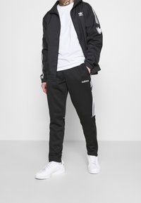 Svart Adidas-träningsjacka med vita ränder, ihop med matchande byxor och en vit t-shirt under. Vita sneakers fulländar outfiten.