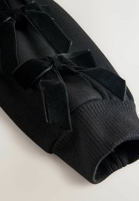 Indumento nero lavorato a maglia con dettagli in nastro di velluto, polsini a coste e una texture liscia. Il design enfatizza linee verticali e bordi netti.