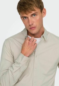 Camisa de botón color beige claro con mangas largas, cuello y textura suave; llevada sobre una camiseta blanca. El modelo ajusta el cuello.