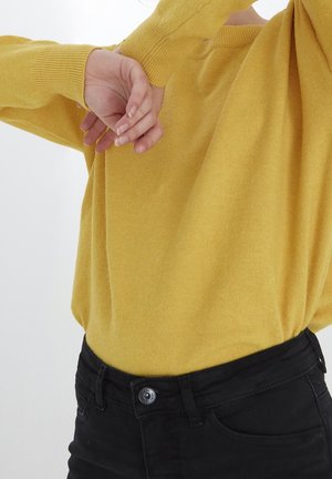 Osoba ubrana w musztardowożółty sweter z długim rękawem i czarne spodnie, z podniesionymi ramionami i rękami z tyłu głowy.