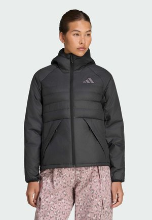 Donna che indossa una giacca nera imbottita con cappuccio e tasche con zip e pantaloni rosa fantasia, che guarda leggermente di lato.