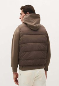 Braune Puffer-Weste mit gesteppten Bereichen, kombiniert mit einem braunen Kapuzenpullover. Verfügt über einen lässigen Schnitt und gerippte Bündchen. Helle Hosen.