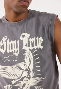 Canottiera grigia in cotone senza maniche con una grande grafica bianca di una figura alata e il testo "Stay True" in un carattere audace.