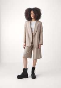 Blazer di lino beige con un solo bottone, abbinato a pantaloni corti beige fino al ginocchio e stivali con tacco neri. Texture liscia, taglio rilassato.