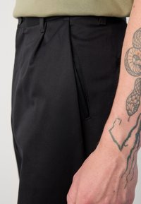 Pantalons noirs sur mesure avec une texture lisse, un devant plissé et des poches latérales. Gros plan sur la ceinture et une main avec un tatouage.