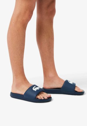 Claquettes en caoutchouc bleu marine avec logo blanc, design à bouts ouverts, et une large bride couvrant le pied, offrant un ajustement décontracté et confortable.