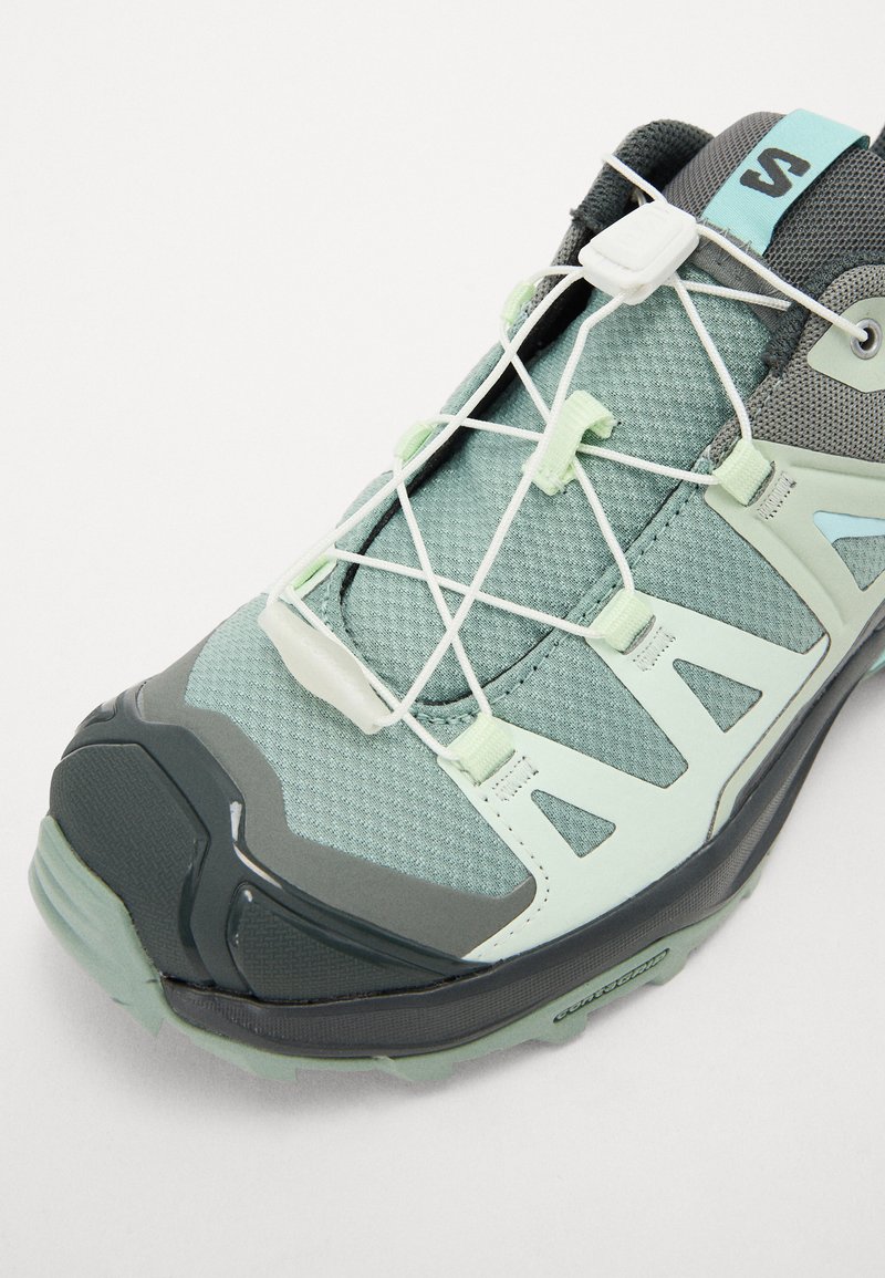 Baskets de sport vert clair et gris avec un dessus texturé, des accents synthétiques et une semelle flexible. Présente un design à lacets avec un système de réglage par curseur.