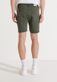 Olijfgroene shorts met een zachte denimtextuur, opgerolde cuffs, twee achterzakken en een logo tag nabij de tailleband. Draagt het in combinatie met witte sneakers.