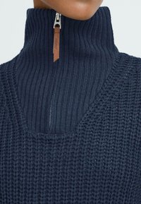 Marineblauwe gebreide trui met een hoge geribbelde kraag, voorzien van een halve rits en leren trekband. Textuurpatroon met een ontspannen pasvorm.