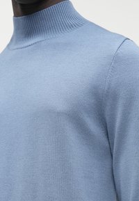 Svetlo modra pletena majica z visokim rebrastim ovratnikom; gladka tekstura in oprijet obliko z minimalnimi šivi za čist videz.