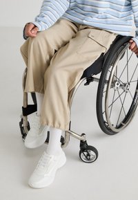 Beige cargo-byxor med vida ben, som bärs tillsammans med vita sneakers och ribbade vita strumpor, av en person i rullstol. Ljust blårandig topp synlig.