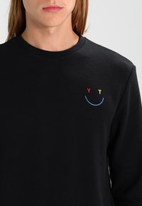 Person som bär en svart sweatshirt med rund halsringning, prydd med en liten broderad leende ansikte gjort av en röd "Y," en gul "T," och en blå kurva på bröstet.