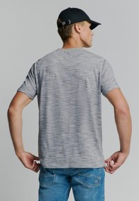 Grijze T-shirt met korte mouwen en een gestructureerd patroon, gecombineerd met lichtblauwe jeans. De persoon draagt een zwarte pet.