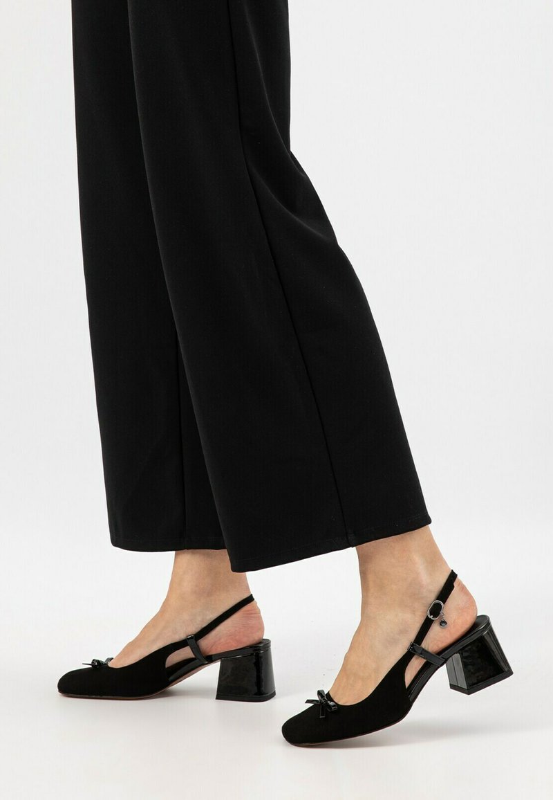 Tacchi slingback neri con un tacco a blocco lucido, parte superiore in morbido camoscio e un dettaglio decorativo a fiocco nella parte anteriore. Cinturino alla caviglia in tessuto piatto.