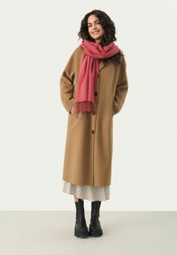 Langer brauner Wollmantel mit schwarzen Knöpfen, kombiniert mit einem pinken Schal und einem hellen Kleid mit Punkten. Schwarze klobige Stiefel runden den Look ab.