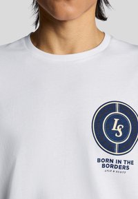 Person iført en hvid T-shirt med et marineblåt cirkellogo og teksten "Born in the Borders Lyle & Scott" på venstre bryst.
