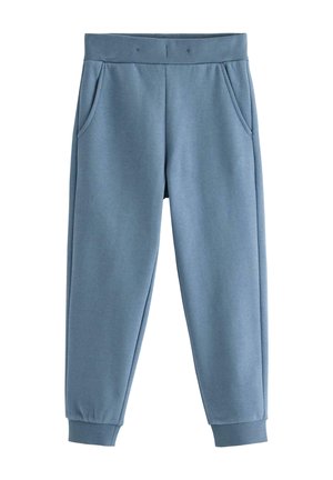 Barn iført blå joggingbukser, navyblå langærmet trøje og hvide og pink sneakers, stående på en lys gulv med en ensfarvet baggrund.