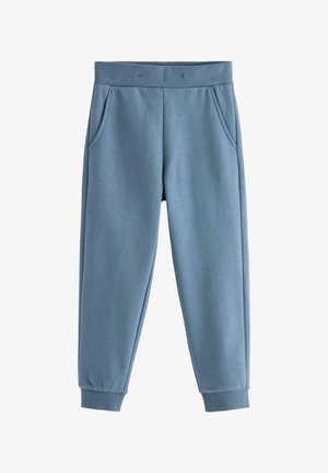 Barn iført blå joggingbukser, navyblå langærmet trøje og hvide og pink sneakers, stående på en lys gulv med en ensfarvet baggrund.