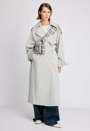 Femme portant un trench-coat gris clair avec un détail d'écharpe à carreaux, un jean large bleu et des chaussures claires, debout devant un fond uni.