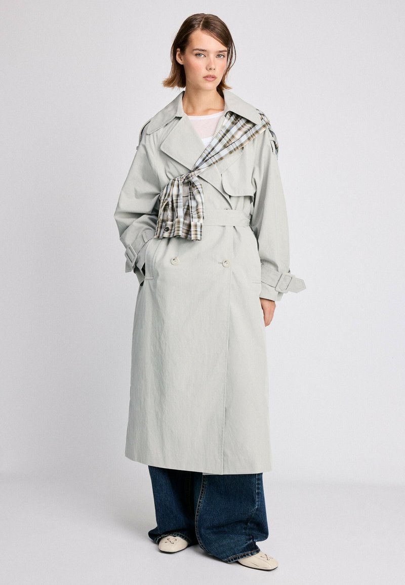 Femme portant un trench-coat gris clair avec un détail d'écharpe à carreaux, un jean large bleu et des chaussures claires, debout devant un fond uni.
