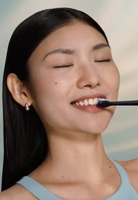 Spazzolino elettrico con manico nero e setole bianche, tenuto da una persona che sorride, indossando una maglietta azzurro chiaro e orecchini di perle.