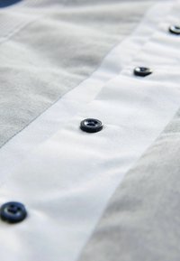 Camisa de tela de algodón que presenta una sección gris y un panel blanco con botones azul oscuro a lo largo de la tapeta para el cierre.