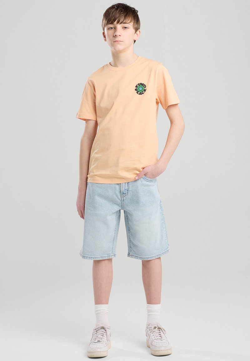 Un ragazzo sta in piedi con sicurezza, indossando una maglietta arancione con una grafica, pantaloni corti azzurri e sneakers bianche, su uno sfondo neutro.