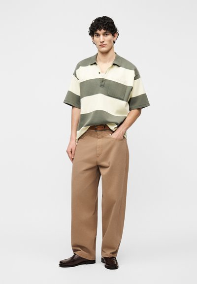 Νεαρός άνδρας στέκεται με το ένα χέρι στην τσέπη, φοράει μπεζ παντελόνι φαρδιάς γραμμής, ριγέ πράσινο και κρεμ πόλο πουκάμισο και σκούρα καφέ παπούτσια.