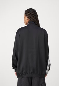 Chaqueta de satén negra con un cuello alto, puños elásticos y dobladillo. Presenta rayas blancas en las mangas. Textura suave, ajuste relajado.