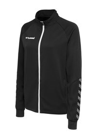 Sort sportjakke med fuld lynlås, hvid lynlås, hvidt "hummel"-logo på brystet og gråt chevronmønster på ærmerne.