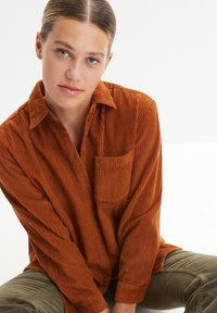 Chemise en velours côtelé couleur rouille avec un col pointu, une poche avant et une texture côtelée verticale. Manches longues et fermeture par boutons.