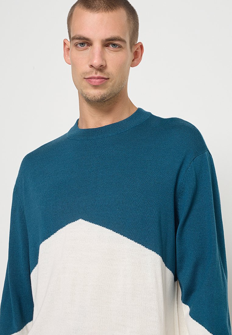 Muškarac s kratkom kosom nosi džemper od pletenine u teal i bijeloj boji, okrenut prema naprijed s neutralnim izrazom lica.