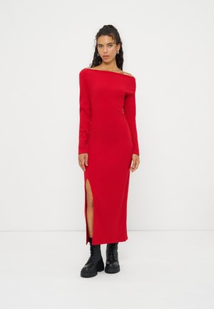 Femme portant une longue robe rouge en maille à épaules dénudées avec une fente sur la cuisse et des bottes épaisses noires à lacets, debout sur un fond blanc.