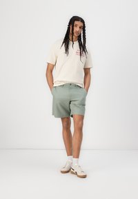 T-shirt crema con grafica rossa, abbinata a pantaloni corti verde chiaro. Il modello indossa sneakers beige con accenti neri e calze bianche.