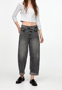 Haut gris à manches longues coupé, associé à un jean gris taille haute à coupe ample avec une finition délavée et des revers roulés. Des mocassins noirs complètent le look.