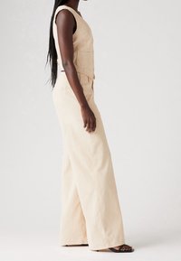 Nainen, jolla on pitkät letitetyt hiukset, on pukeutunut heti hihattomaan beigeen liiviin ja leveälahkeisiin housuihin sekä mustiin korkokenkiin, seisten sivuttain yksinkertaista taustaa vasten.