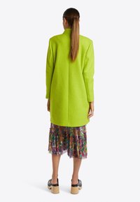 Bright green textured coat with a relaxed fit, paired with a multi-colored floral sheer skirt and clogs. Back view shows the coat's length.