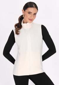 Vest din fleece cu fermoar, de culoare crem, cu guler înalt și fermoar alb, asortat cu un top negru cu mânecă lungă. Textură netedă, croială ajustată.