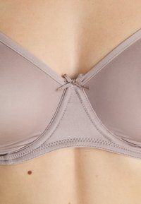 Light taupe bra with soft fabric, padded cups, and a triangle shape. Features a small sewn bow at the center and smooth stitching along edges.