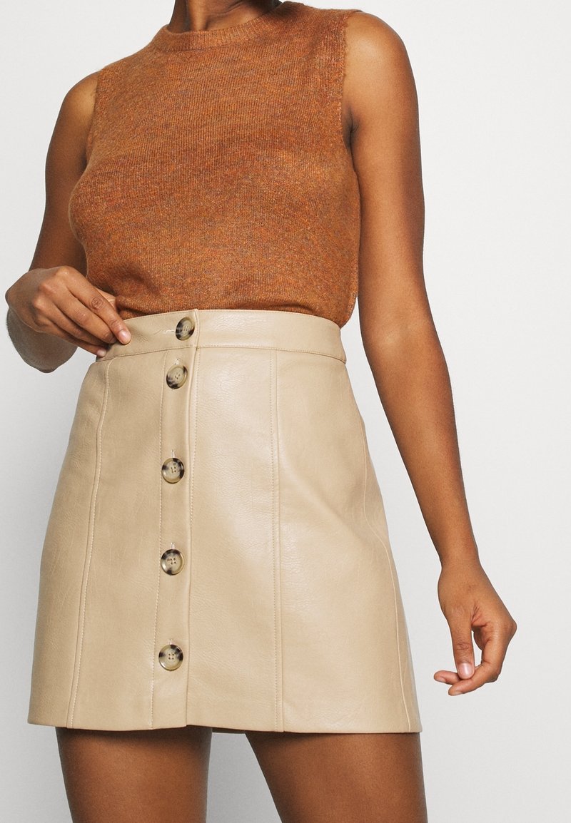 Femme portant un haut sans manches en maille marron associé à une jupe en cuir beige taille haute avec une fermeture à boutons sur le devant.