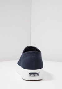 Námořnické plátěné tenisky s bílou gumovou podrážkou, zobrazené zezadu s logem značky "Superga" na patě.