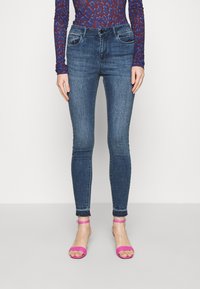 Jeans skinny de denim azul oscuro, con cintura de altura media, bolsillos frontales y dobladillos deshilachados, acompañados de sandalias rosas con tiras.
