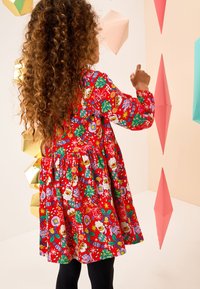 Robe rouge ornée de motifs festifs de Noël, incluant le Père Noël, des biscuits en pain d'épice et des éléments décoratifs. Manches longues, jupe froncée.