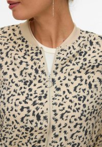 Sieviete valkā bēša un melnas krāsas rakstainu rāvējslēdzēja jaku ar riņķveida rāvējslēdzēja svilpi un garu, smalku ķēžu auskaru.