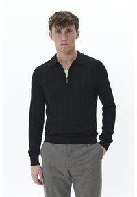 Sudadera negra con cremallera, fabricada en un tejido texturizado, con puños y cuello acanalados. Combinada con pantalones gris a rayas. El modelo está de pie contra un fondo blanco.