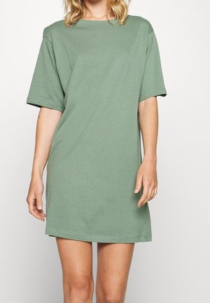 Kobieta ubrana w luźną sukienkę T-shirt w kolorze szałwii z krótkim rękawem, stojąca na jednolitym białym tle.